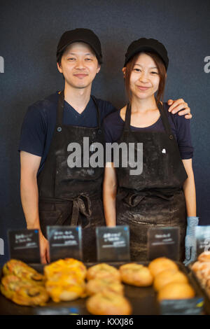 Smiling man and woman wearing baseball cap et tablier, debout dans une boulangerie, des plateaux avec des marchandises. Banque D'Images