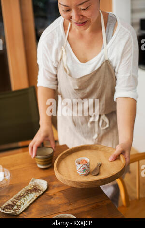 Smiling woman wearing apron holding petit plateau de bois rond avec bol de gingembre écrasé et cuillère, tasse mise sur table. Banque D'Images