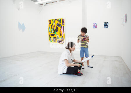 Femme aux cheveux noirs portant une chemise blanche assis sur le plancher en galerie d'art avec un stylo et du papier, garçon debout à côté d'elle. Banque D'Images