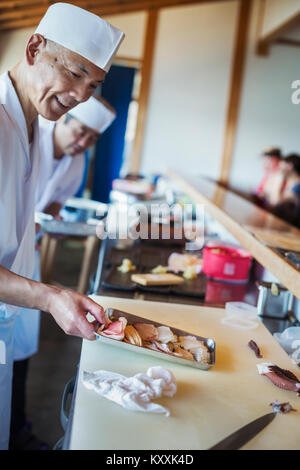 Deux chefs travaillant au comptoir dans un restaurant de sushi japonais, tenant le plateau métallique avec du poisson et des fruits de mer. Banque D'Images