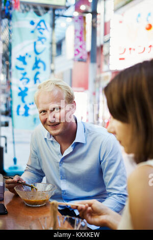 Young man smiling at camera et femme assise à une table dans un restaurant fast food asiatique, à l'aide de baguettes, mange des nouilles à partir d'un bol en verre. Banque D'Images