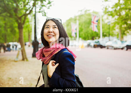 Femme souriante aux cheveux noirs portant veste bleu et foulard rouge debout sur le côté d'une route bordée d'urban. Banque D'Images