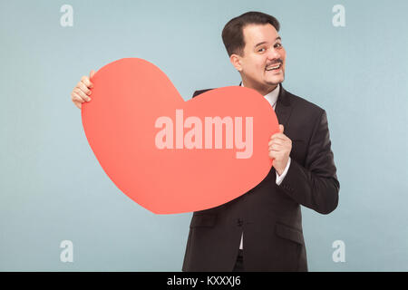 Bearded businessman holding big red heart shape et souriant à pleines dents. Studio shot, isolé sur fond bleu clair Banque D'Images