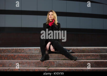 Fashion femme blonde en manteau noir assis sur les marches Banque D'Images