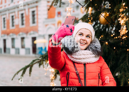 Riga, Lettonie. Belle Jeune Fille Pretty Caucasian Woman Dressed In White Hat et Red Jacket s'amusant et en prenant des photos sur Smartphone Selfies sur Retour Banque D'Images