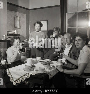 Années 1950, historiques, des familles heureuses dans la Grande-Bretagne d'après-guerre... nous voyons ici une famille bénéficiant d'une bonne tasse de thé ensemble autour de la table de cuisine, avec le bonus de biscuits et muffins. Banque D'Images