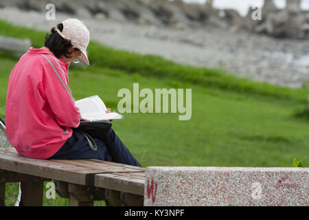 Femelle adulte avec képi siège au banc en bois et lit un livre au parc au bord de mer, de la ville de Hualien, Taiwan Hualien County, Banque D'Images