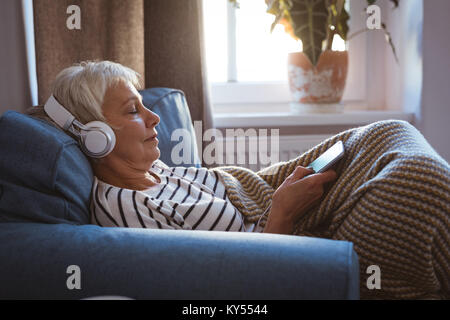 Senior woman liste pour la musique sur téléphone portable assis sur un canapé dans la salle de séjour Banque D'Images