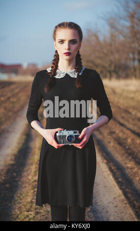 Outdoor portrait of a cute jeune fille en robe noire à l'ancienne avec caméra film vintage en mains Banque D'Images