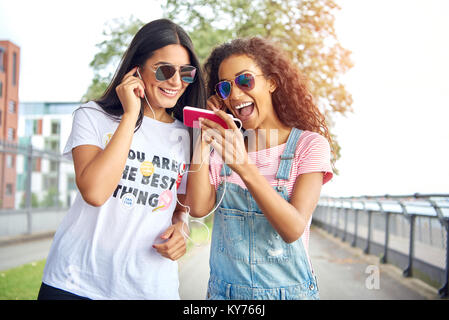 Deux jeunes copines rire marcher dans une promenade dans l'été à l'écoute de la musique ensemble sur une paire d'écouteurs Banque D'Images