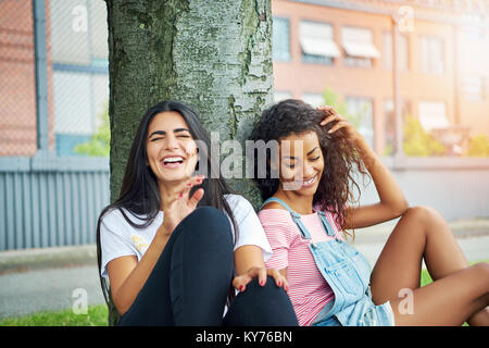 Deux jeunes amies de rire tandis qu'assis sur une pelouse, se penchant en arrière sur un tronc d'arbre sur une journée ensoleillée Banque D'Images