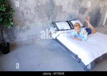 Jolie jeune femme jouit de confort de la chambre et se détend, compose le numéro de téléphone mobile et communique avec petite amie, allongée sur un grand lit avec des draps blancs Banque D'Images