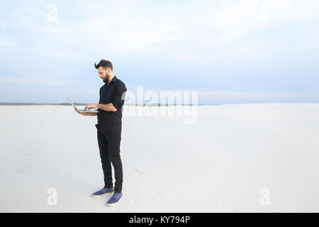 Auteur avec portable, rendez-vous à mer, le jeune homme s'inspirer pour réserver des voyages. Barbu en chemise noire des sables bitumineux de regarder autour de soi. Concept Banque D'Images
