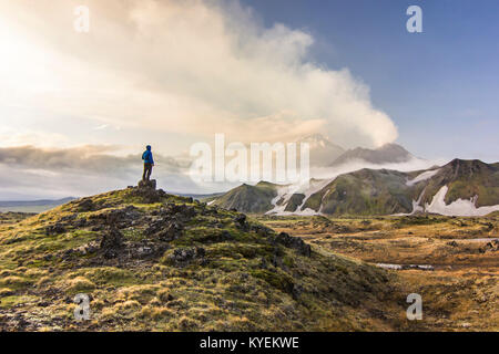 L'homme à veste bleue debout sur champ vert près de volcans en Bezymyannyi et Kamen', du Kamtchatka avec collines enneigées au coucher du soleil Banque D'Images