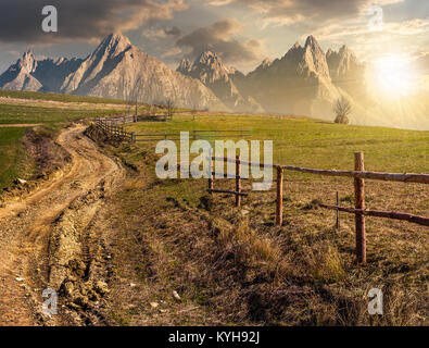 Route à travers les champs en milieu rural région montagneuse. imagerie composite de la campagne agricole de printemps au coucher du soleil. clôture en bois le long de l'herbe des fi Banque D'Images