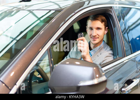 Portrait de beau jeune homme assis à l'intérieur de voiture de luxe et holding car keys souriant joyeusement à caméra dans showroom Banque D'Images