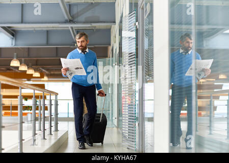 Portrait of handsome man with suitcase walking in corridor d'un établissement moderne, lisant le journal sur le chemin Banque D'Images