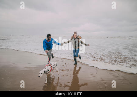 Un jeune couple de fuir les vagues en marchant leur chien sur une plage d'hiver. Banque D'Images