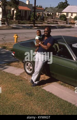 Portrait d'un homme posant avec un bébé garçon dans ses bras, comme il s'appuie contre une voiture garée dans l'entrée d'un quartier résidentiel de Los Angeles, Californie, juillet 1975. () Banque D'Images