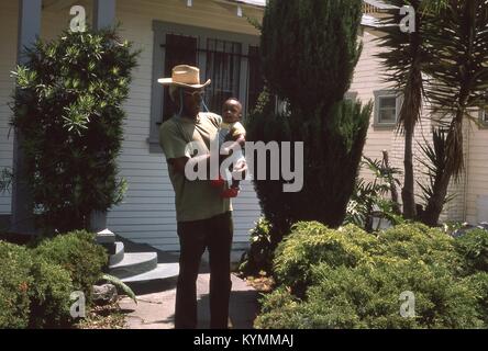 Portrait of a smiling African American man un nourrisson African American boy, à l'extérieur d'une maison à Los Angeles, Californie, juillet 1975. L'homme porte un chapeau de paille et lunettes cerclées d'or, et le bébé porte des bretelles et de tricot rouge booties. () Banque D'Images