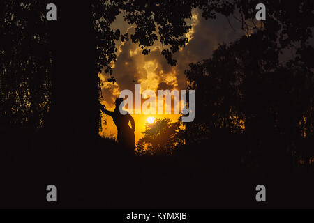 Silhouette d'une femme appuyée contre big tree, jaune coucher du soleil illuminant ciel derrière sa Banque D'Images