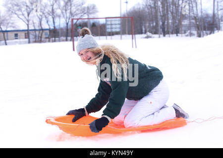 Un rire adolescente en pantalon de ski et neige chapeau sur ses longs cheveux glissant sur une colline sur un toboggan dans un paysage d'hiver Banque D'Images