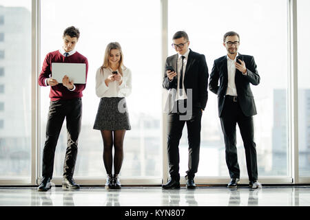 D'affaires du groupe permanent d'amis et à l'aide de gadgets modernes contre l'arrière-plan de windows. Banque D'Images