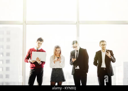 D'affaires du groupe permanent d'amis et à l'aide de gadgets modernes contre l'arrière-plan de windows. Banque D'Images