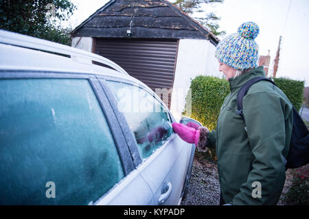 White middle-aged woman dans un bobble hat avec un grattoir mitaine rose efface les glaces au large de sa voiture avant d'argent qu'elle va travailler à un matin d'hiver. Banque D'Images