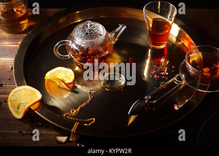 Petite théière en verre chaud avec du thé noir, tasse, des verres, des pétales de rose, loupe de poche sur la chaîne d'or, tranche d'orange sur le plateau d'or sur bois Banque D'Images