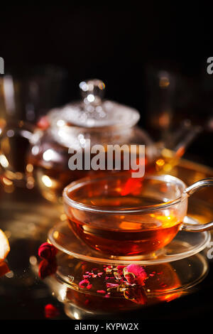 Gros plan vertical de petite théière en verre et tasse à thé rouge chaud, les pétales de rose, loupe de poche sur chaîne dorée sur bac d'or soir lig Banque D'Images