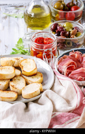 Ingrédients pour faire des tapas ou bruschetta. Pain croustillant, jambon prosciutto, tomates séchées, huile d'olive, les olives, le poivre, les verts sur les plaques sur fond blanc p Banque D'Images