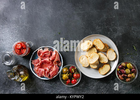 Ingrédients pour faire des tapas ou bruschetta. Pain croustillant, jambon prosciutto, tomates séchées, huile d'olive, les olives, le poivre, les verts sur les plaques sur te foncé Banque D'Images