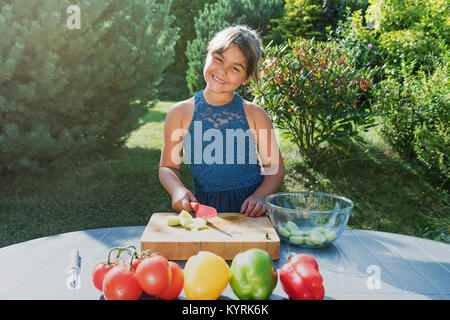 Smiling little girl est salade cuisson de légumes frais à l'extérieur. Jeune fille est à la recherche de l'appareil photo. Banque D'Images