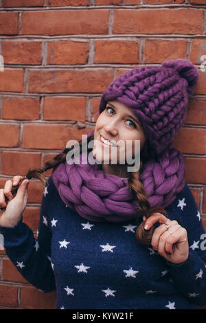 Brunette woman in hat tricoté violet, en l'infini d'écharpe et pull bleu à étoiles blanches sur elle faire posant avec mur de brique sur l'arrière-plan Banque D'Images