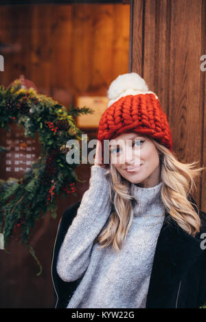 Young blonde woman in red hat tricoté avec pompon blanc, gris pull, veste noire et un jean bleu faire posant avec Christmac cafe fenêtre sur le backg Banque D'Images