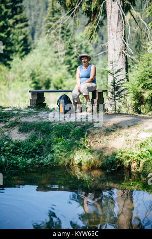 Femme assise sur le banc des forêts en vertu de l'arbre, près de l'eau. Portrait dame d'âge moyen se reposer après une randonnée dans la nature sur l'été chaude journée. Paysage pittoresque Banque D'Images