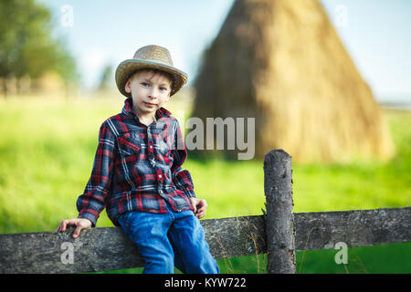 Siège enfant sur clôture en bois contre la botte de pittoresque, Close up. Jeune garçon dans les tenues de se reposer sur une clôture en village. Country Vacations concept Banque D'Images