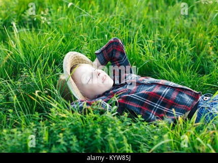 Joli petit garçon couché sur l'herbe sur le dos, les mains derrière la tête. Peu rêveur se trouve dans la région de prairie avec chapeau, les yeux fermés. Enfant endormi et souriant en natu Banque D'Images