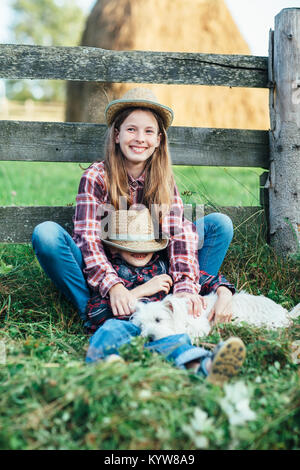 Sœur serrant avec frère assis avec chien près de clôture en bois contre de haystack. Enfance heureuse vacances. Drôle de famille membres le village. Détendez-vous c Banque D'Images