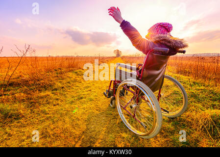 Femme heureuse au coucher du soleil. Une jeune fille dans un fauteuil roulant - vue arrière Banque D'Images