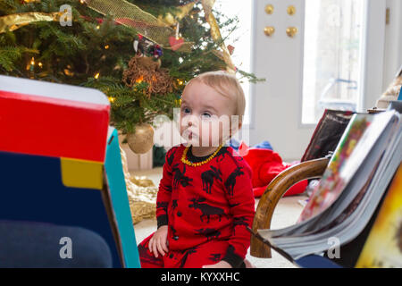 Triste 1 ans enfant en pyjama rouge assis sur le plancher à côté de l'arbre de Noël Banque D'Images