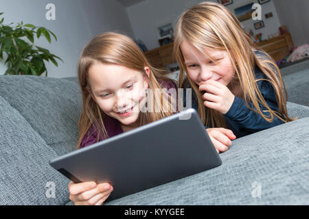 2 filles se coucher sur le canapé et regarder joyeusement à l'écran d'une tablette Banque D'Images