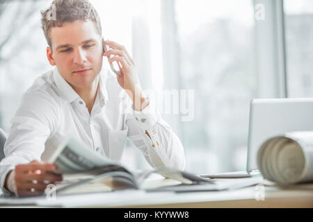 Jeune businessman reading brochure using cell phone at office desk Banque D'Images