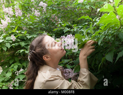 Jeune fille au manteau frisé beige est debout près de lilas en fleurs et de fleurs. L'inhalation de Bush Banque D'Images