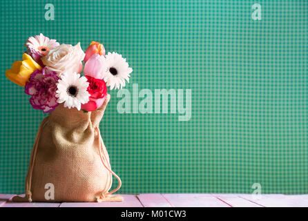 Bouquet de gerberas multicolores, tulipes et roses dans un joli sac en toile de jute, sur une table en bois de rose, et un fond en tissu vert, en lumière naturelle. Banque D'Images