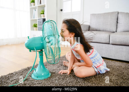 Jolie jolie petite fille assise sur le plancher du salon jouant le ventilateur électrique et l'expression orale se sentir drôle, durant la saison estivale. Banque D'Images