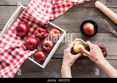 Pommes rouges bien mûrs dans une boîte de bouleau sur une planche de bois. Femme éplucher les pommes pour la cuisine un couteau spécial, des ustensiles de cuisine. Style rustique. Vue d'en haut. Banque D'Images