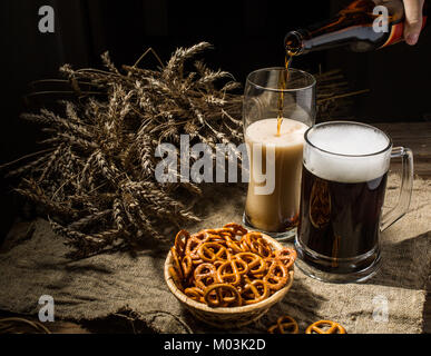 Bouteille de bière mousseuse versée dans glasse avec mug permanent de la bière avec du blé et panier de bretzels Banque D'Images