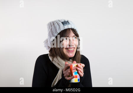 Portrait d'une femme heureuse en intérieur, chapeau et écharpe en laine, mains froides sur une tasse de thé et de café. Un sourire authentique et naturel, agréable, grand et gloueux Banque D'Images